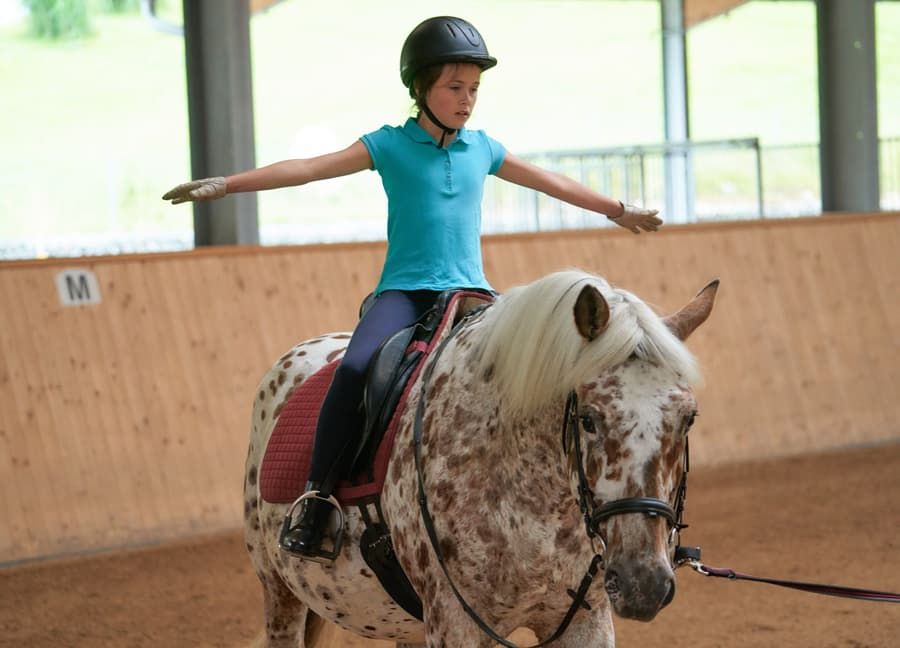 Kind balanciert mit ausgestreckten Armen auf einem Pony in der Reithalle des Familienhotels Schreinerhof